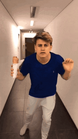 a young man in a blue shirt and white pants is walking down a hallway holding a cup