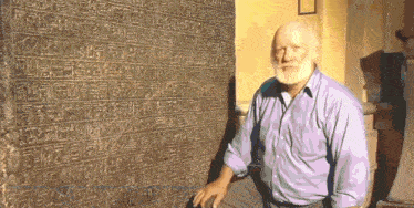 a man in a purple shirt sits in front of a large stone wall with arabic writing on it