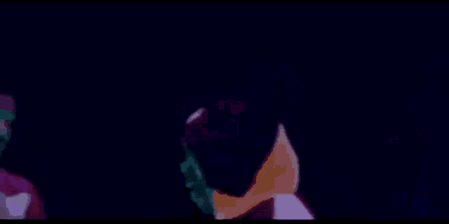 two men are standing next to each other in a dark room . one of the men is wearing sunglasses and a hat .