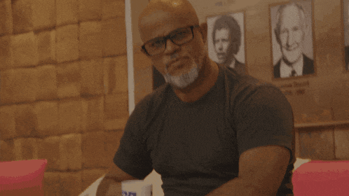 a bald man with glasses and a beard stands in front of a wall with pictures of famous people including tony blair