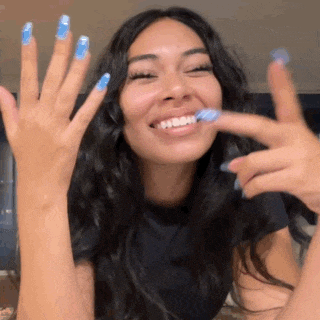 a woman with long black hair and blue nails is smiling and holding up her hands .