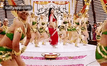 a woman in a red saree is dancing in front of a group of dancers in green and gold costumes .