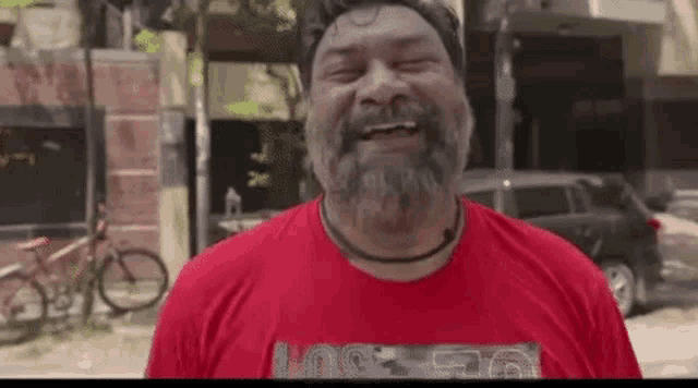 a man with a beard wearing a red t-shirt is smiling with his eyes closed .