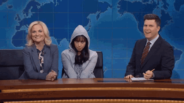 a woman wearing a hoodie sits at a desk with two news anchors