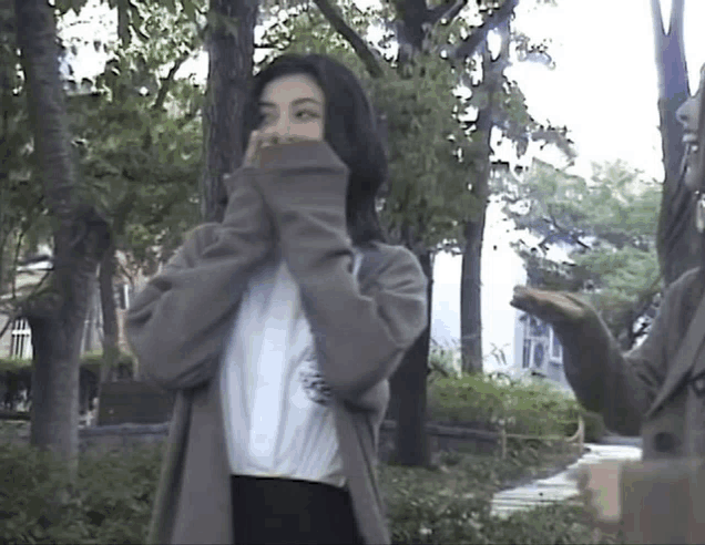 a woman is covering her face with her sweater while standing in a park with trees in the background .