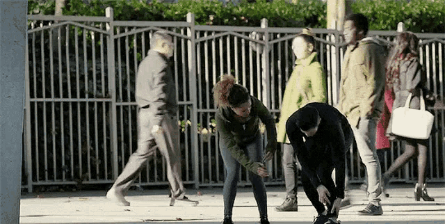 a man and a woman are tying their shoes on a sidewalk while people walk by .