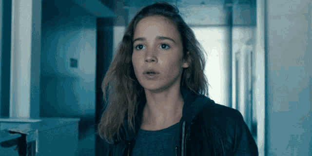 a woman in a black jacket stands in a hallway looking at the camera