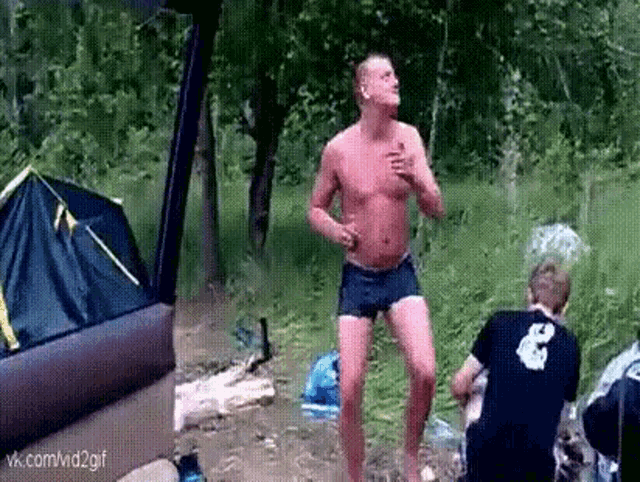 a man without a shirt is dancing in the woods while another man watches .