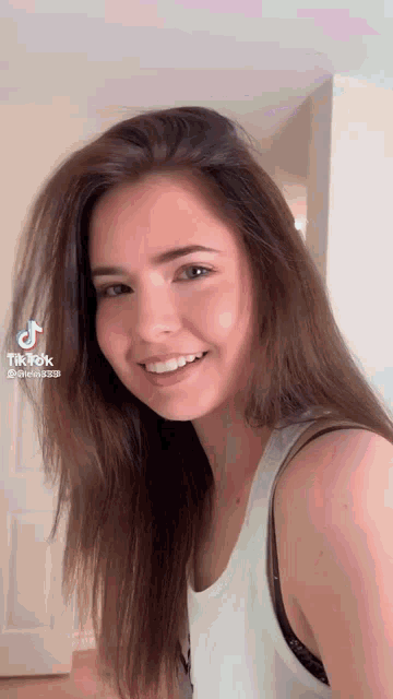 a woman with long brown hair is smiling and wearing a white tank top and black bra .