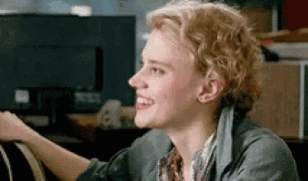 a woman with curly blonde hair is smiling while sitting at a table in front of a computer monitor .