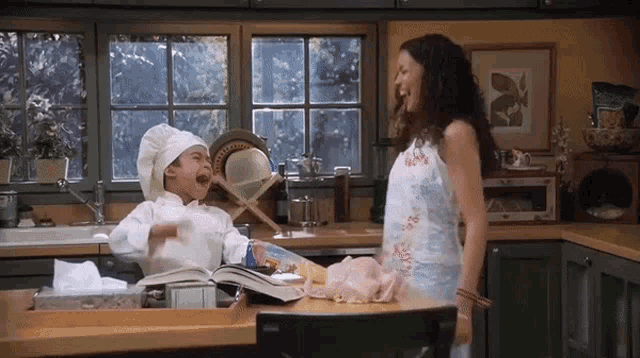 a woman laughs while a little boy in a chef 's hat sits at a counter
