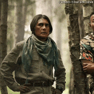 a man wearing a scarf and a watch stands in the woods with ramon-tikaram-love written above him