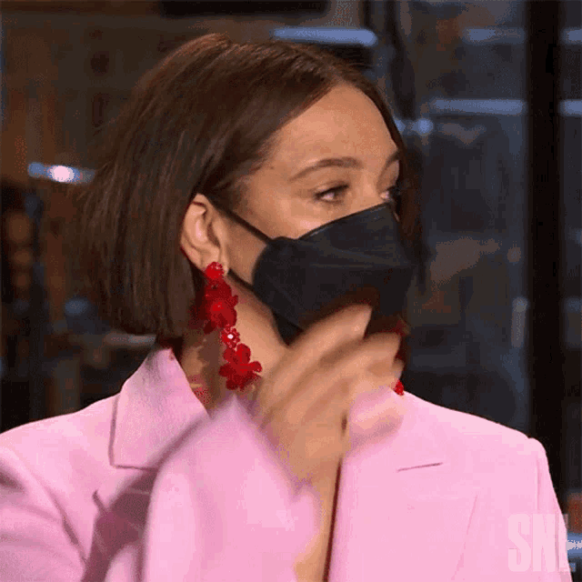 a woman wearing a pink jacket and red earrings is wearing a face mask .
