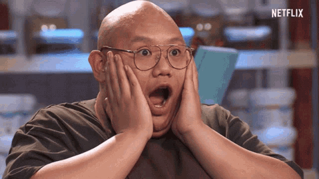 a bald man with glasses is making a surprised face while holding his hands to his face .