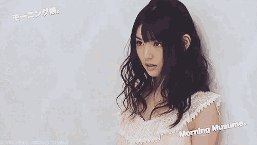 a woman in a white dress is standing in front of a wall and looking at the camera .