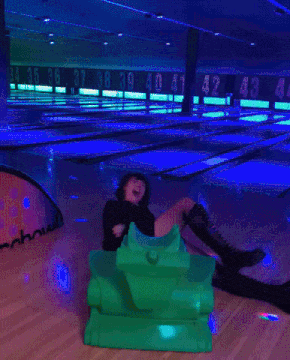 a woman is sitting on a green toy tank in a bowling alley