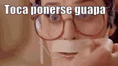 a woman wearing glasses is holding a piece of tape over her mouth and says toca ponerse guapa .