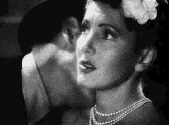 a woman wearing a pearl necklace and a flower in her hair is whispering into another woman 's ear .