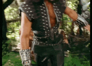 a man is standing in the woods wearing a leather vest and gloves .