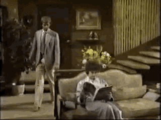a man and woman are standing in a living room while a woman sits on a couch reading a book .