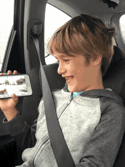 a young boy is sitting in a car seat holding a cell phone with a picture of a car on it