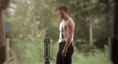 a man in a white tank top and suspenders is standing in front of a fence