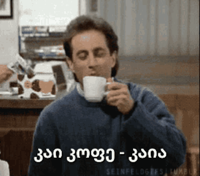 a man in a blue sweater drinking from a white cup with the words seinfeld on the bottom