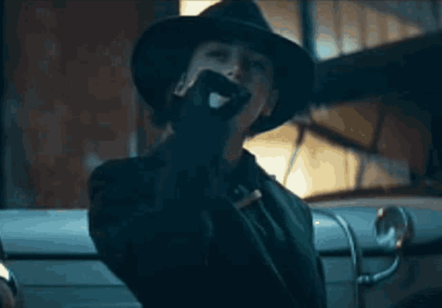 a woman wearing a hat and gloves is smoking a cigarette in front of a car .