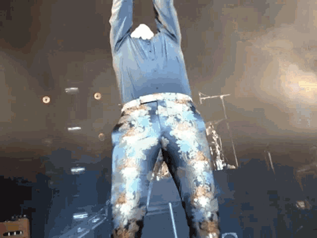 a man in a blue shirt and floral jeans is standing on a stage with his arms in the air