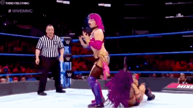a woman with pink hair is wrestling another woman in a wrestling ring while a referee looks on .