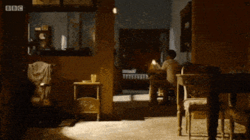 a man in a white t-shirt is sitting on a couch in a dark room holding a book .