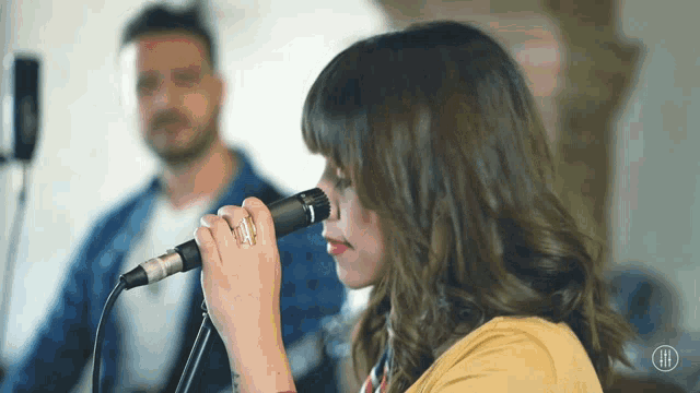 a woman singing into a microphone with a man playing a guitar behind her