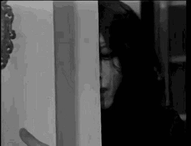a black and white photo of a woman peeking out of a door .