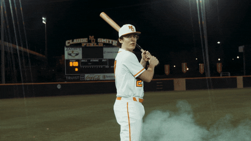 a baseball player is holding a bat in front of a scoreboard that says claude m smith field