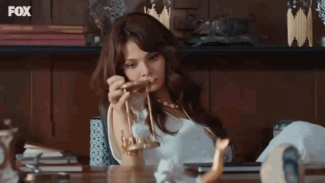 a woman is sitting at a desk holding a hourglass in front of a fox logo .