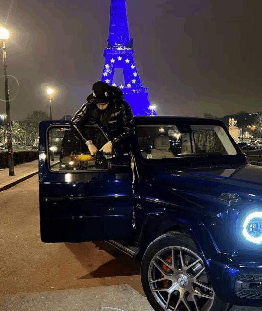 a man in a black jacket is sitting in a blue car in front of a blue eiffel tower