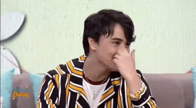a young man in a striped shirt is sitting on a couch covering his mouth with his hand .