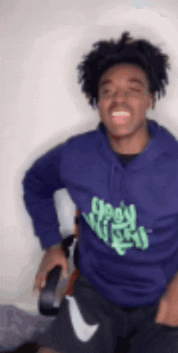 a young man wearing a purple hoodie and black shorts is standing in front of a white wall and smiling .
