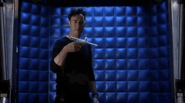 a man in a black shirt holds a gun in front of a blue wall