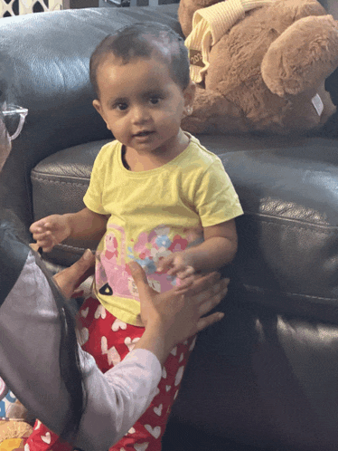 a little girl in a yellow shirt is sitting on a couch next to a teddy bear