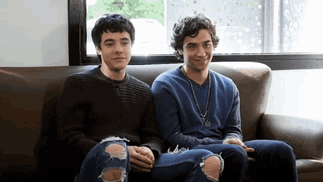 two young men sit on a couch holding hands
