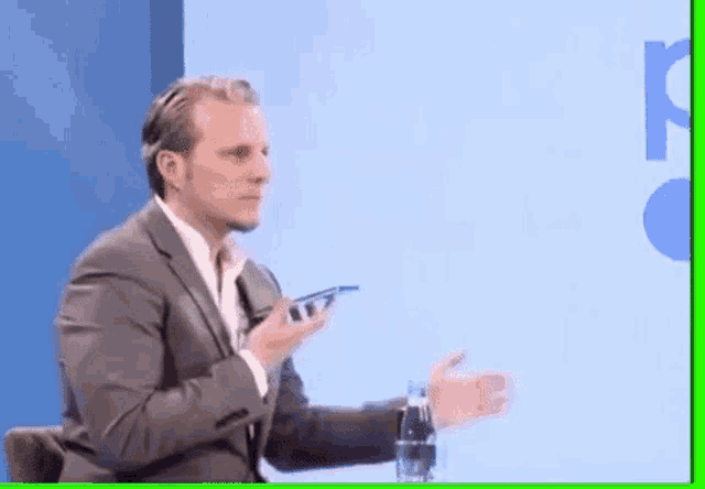 a man in a suit and tie is sitting in front of a blue wall holding a cell phone .