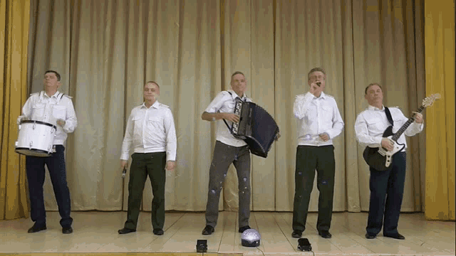 a group of men are standing on a stage playing instruments including a drum and a guitar