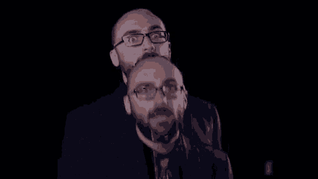 a man with glasses and a beard is holding another man 's head in a dark room .