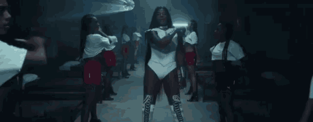 a woman in a white bodysuit is standing in front of a group of women in red shorts .