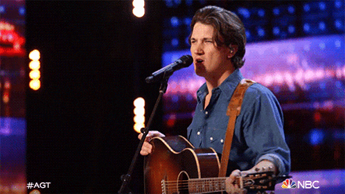 a man singing into a microphone while holding a guitar in front of a nbc logo