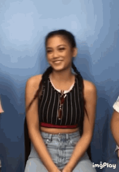 a woman wearing a striped crop top and jeans is smiling while sitting in front of a blue wall .