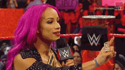 a woman with pink hair is standing in a wrestling ring talking into a microphone .