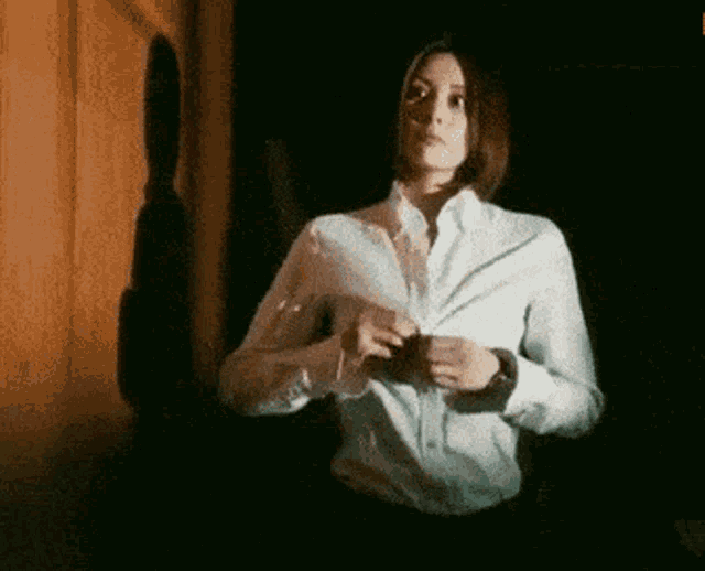 a woman in a white shirt is standing in a dark room taking off her shirt .