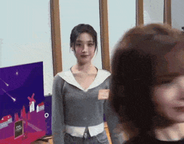 a woman in a gray sweater is standing next to another woman in front of a television .